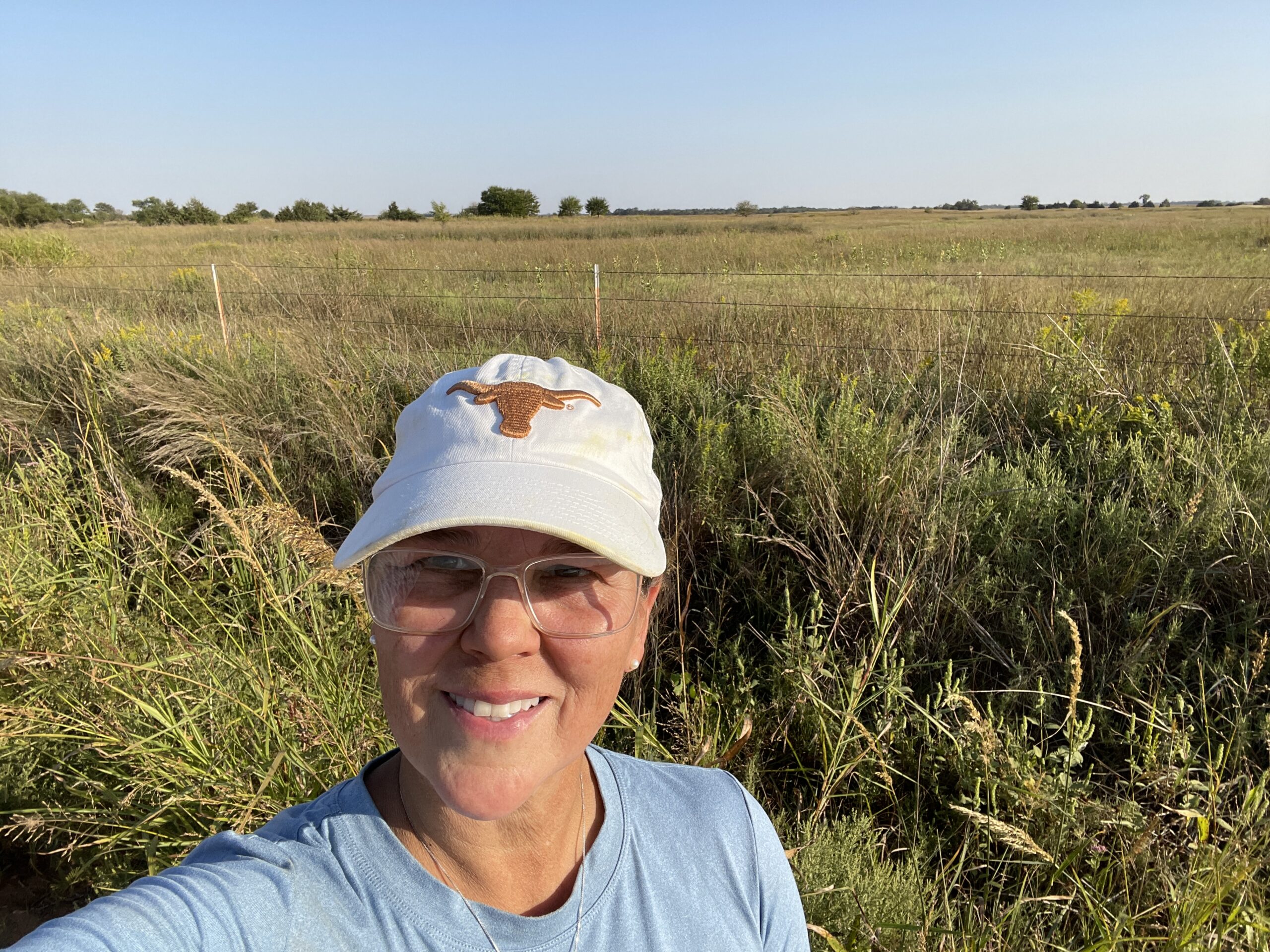 Meg Laakso in a ball cap and glasses, smiling, with the farm's grassland and fence line stretching out behind her